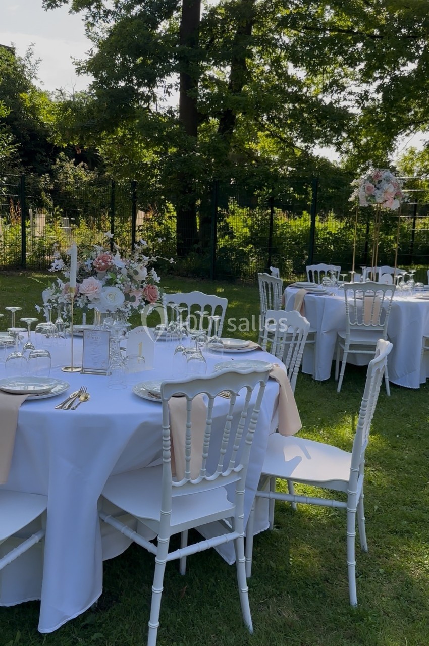 Tables rondes décorées avec nappes blanches, fleurs et chaises blanches, disposées dans un jardin verdoyant.