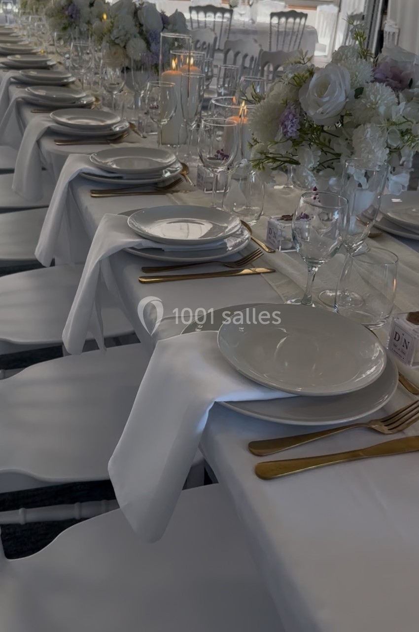 Tables dressées avec nappes blanches, vaisselle élégante, couverts dorés et centres de table fleuris dans une salle…