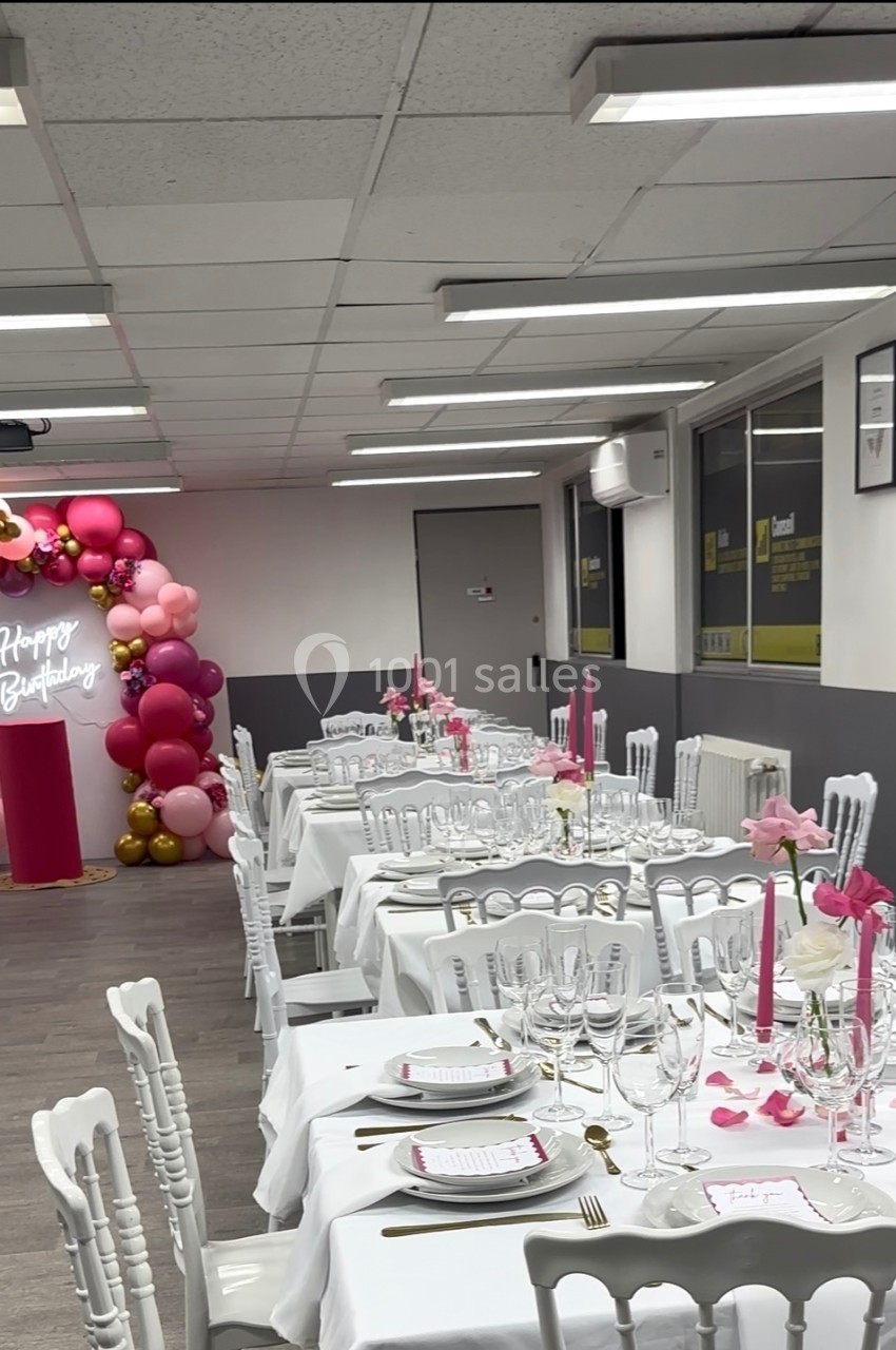 Salle décorée pour un anniversaire avec tables dressées, chaises blanches et arche de ballons roses et dorés.