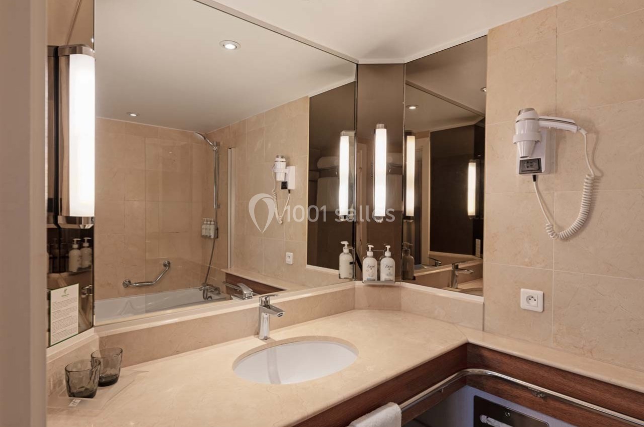 Salle de bain avec lavabo, miroir, baignoire équipée d'une barre d'appui, sèche-cheveux mural et éclairage intégré.