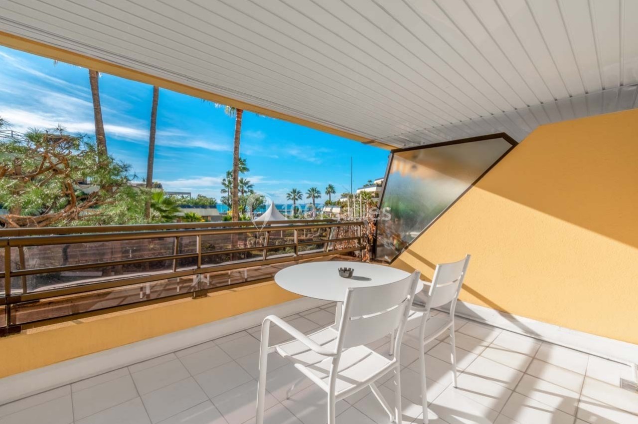 Balcon ensoleillé avec table et chaises blanches, vue sur des palmiers, la mer et un ciel bleu dégagé.