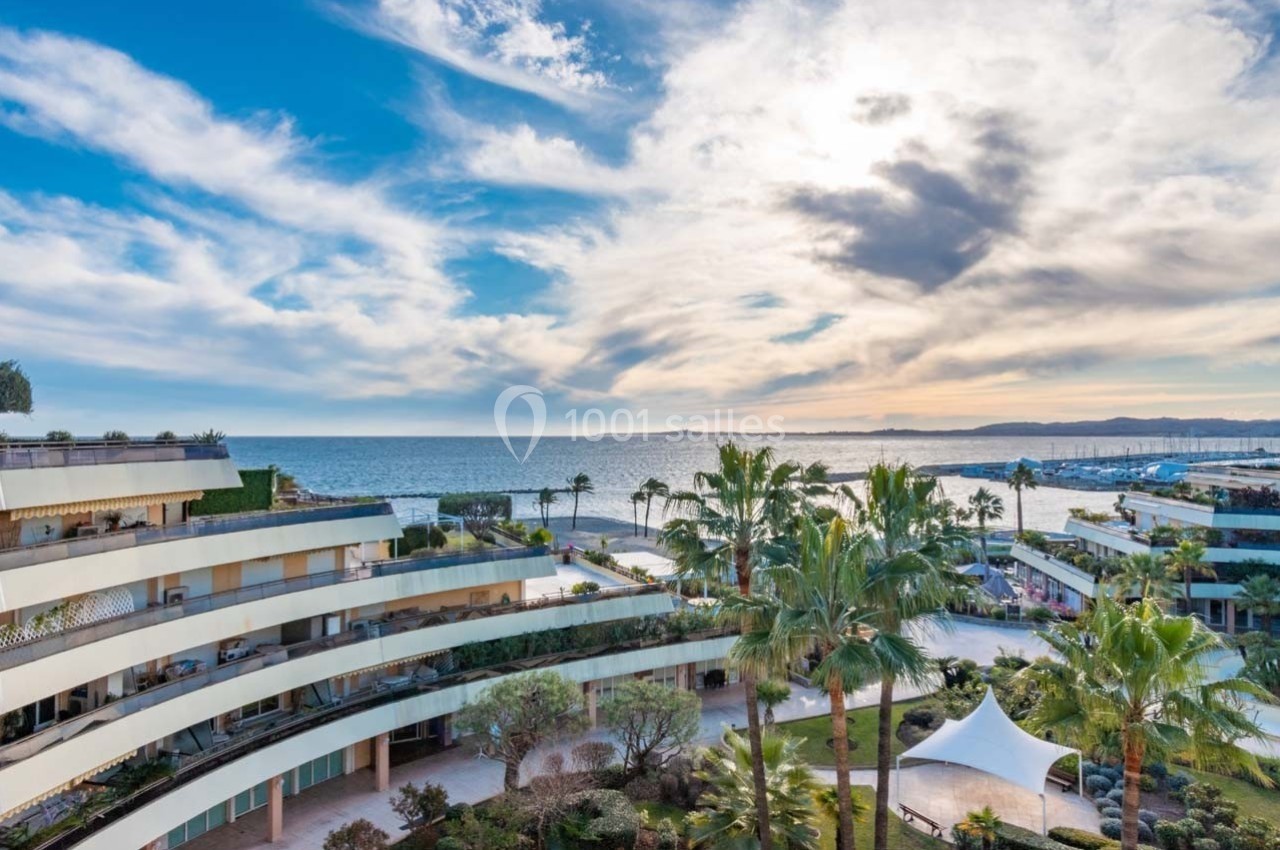 Vue sur des bâtiments modernes entourés de palmiers avec la mer et un ciel partiellement nuageux en arrière-plan.