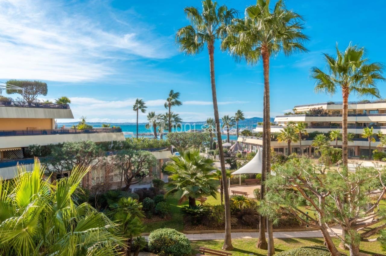 Vue sur un jardin arboré avec palmiers, bâtiments modernes et la mer en arrière-plan sous un ciel dégagé.