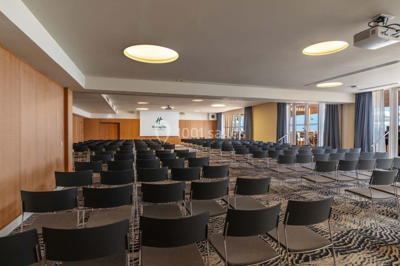 Salle de conférence avec rangées de chaises noires, éclairage moderne et écran de projection au fond.
