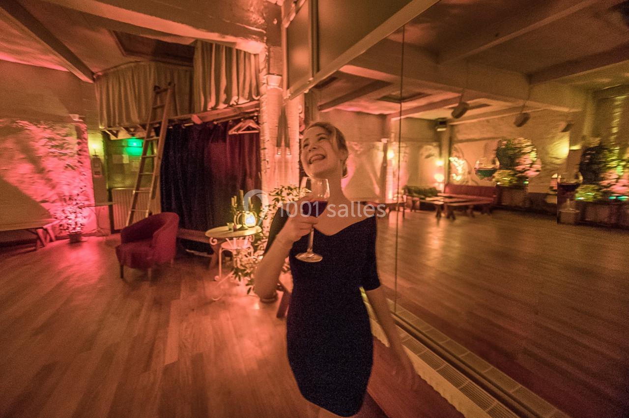 Femme souriante tenant un verre de vin dans une salle chaleureusement éclairée avec des miroirs et des fauteuils.