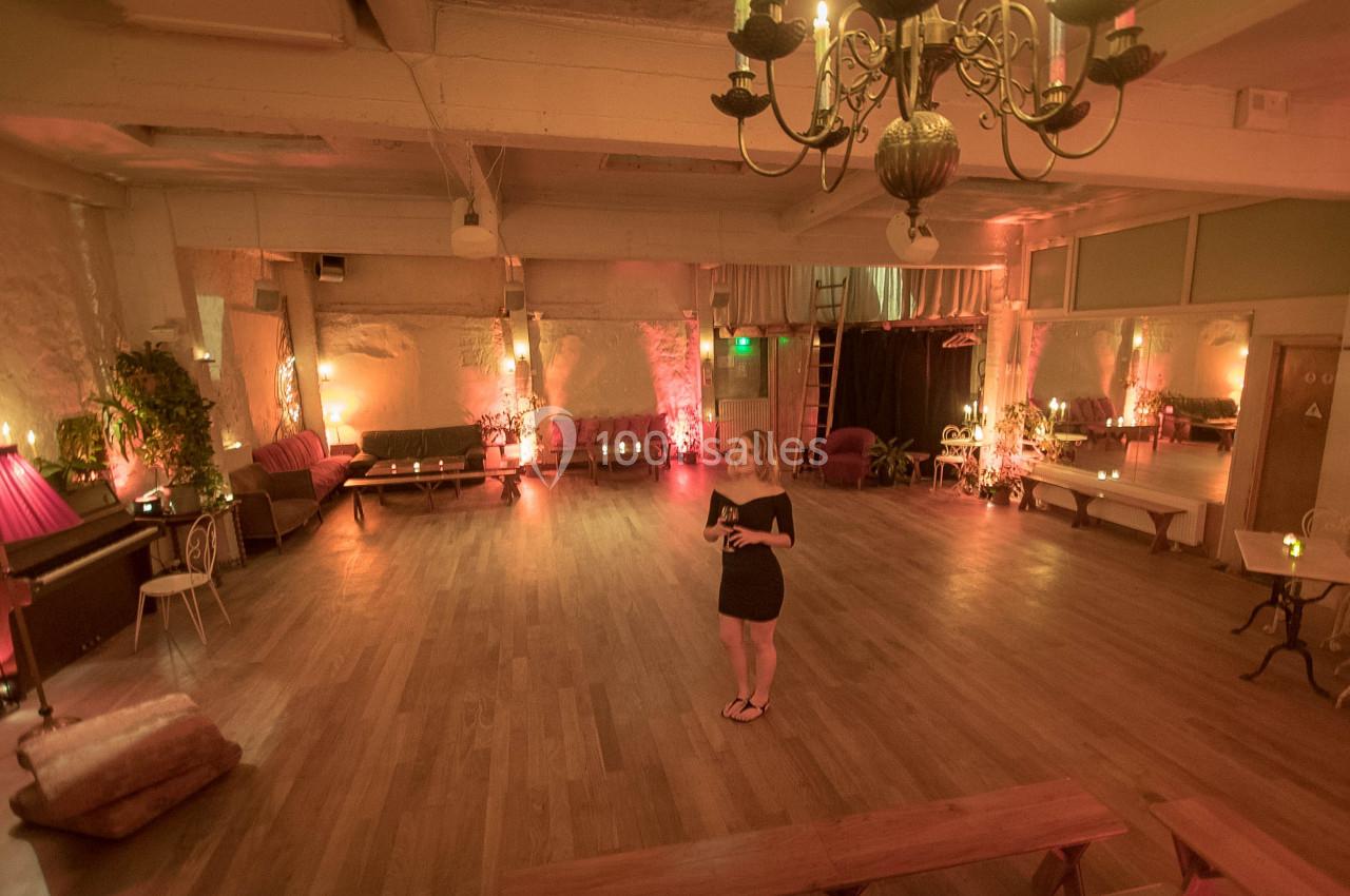 Une femme se tient au centre d'une grande salle chaleureusement éclairée, avec parquet, canapés et lustre suspendu.