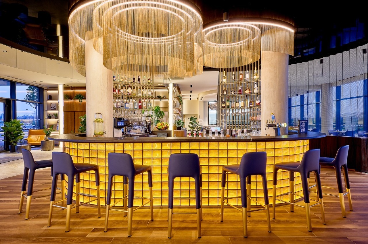 Bar moderne avec comptoir lumineux en verre, tabourets bleus et dorés, et lustres circulaires suspendus.