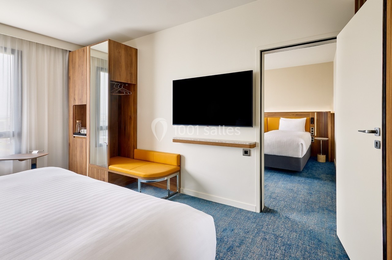 Chambre d'hôtel moderne avec lit, télévision murale, espace de rangement en bois et vue sur une seconde chambre.