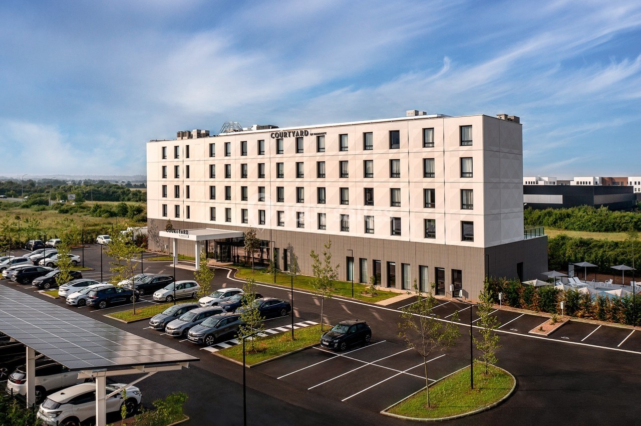 Façade d'un hôtel moderne avec parking extérieur, entouré de végétation et sous un ciel dégagé.