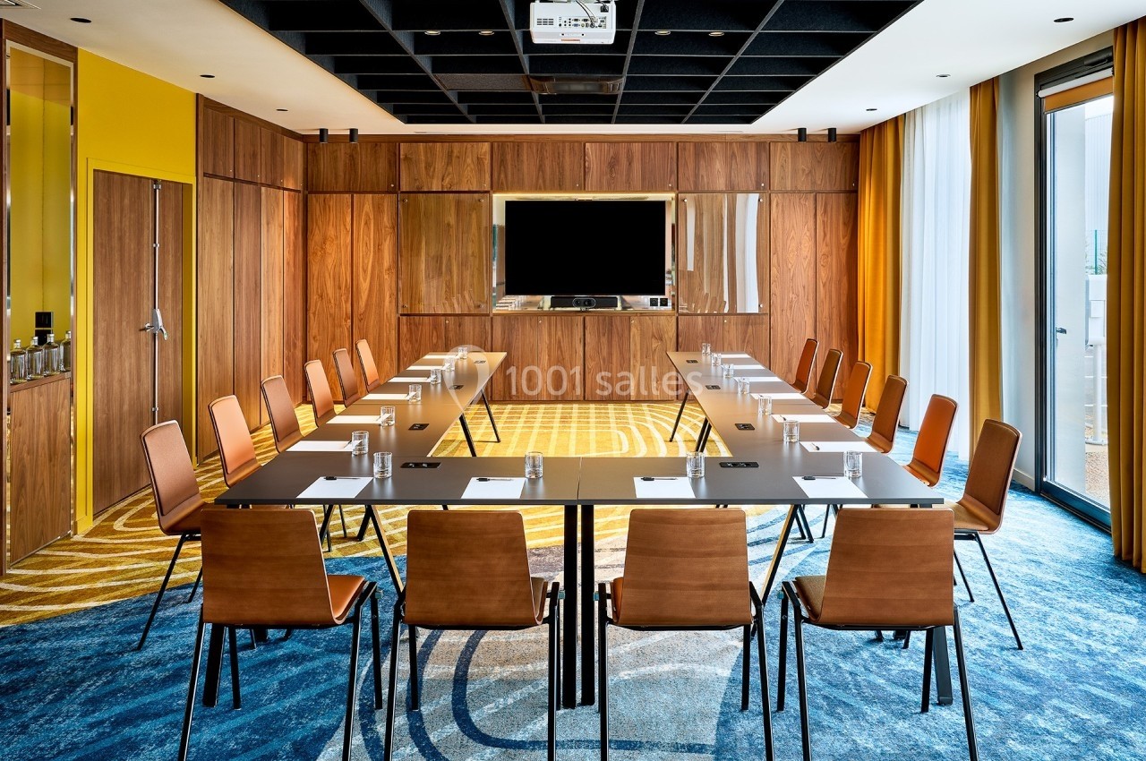 Salle de réunion moderne avec table en U, chaises en bois, écran mural et grandes fenêtres laissant entrer la lumière…