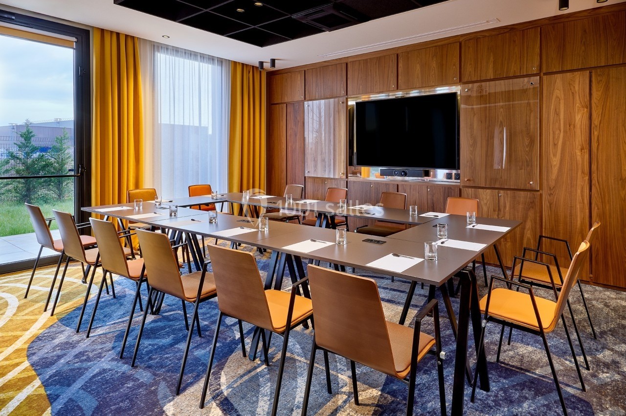 Salle de réunion moderne avec table rectangulaire, chaises en bois, écran mural et grande fenêtre laissant entrer la lumière…