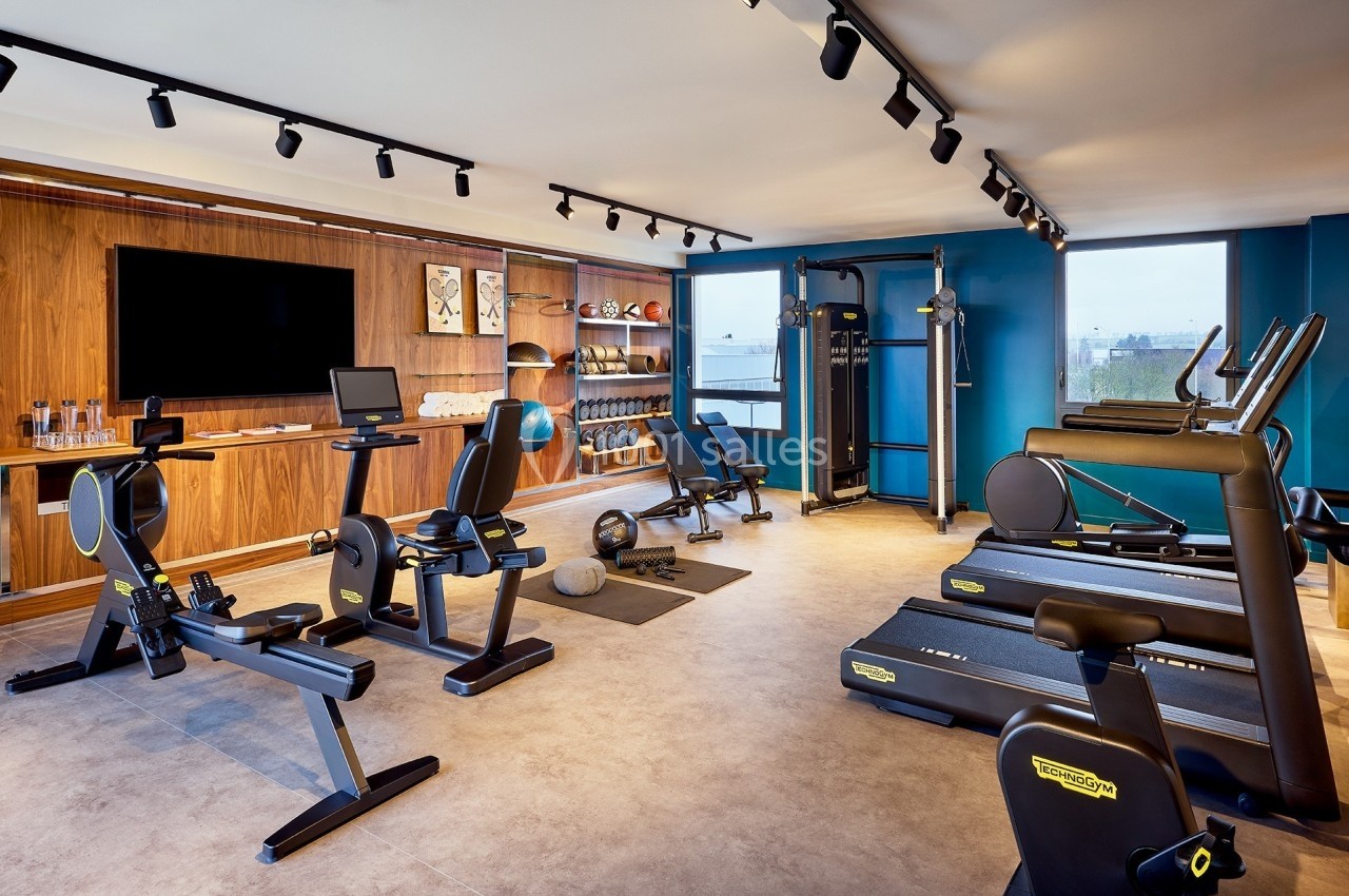 Salle de sport moderne équipée de tapis de course, vélos, rameurs, appareils de musculation et écran mural.