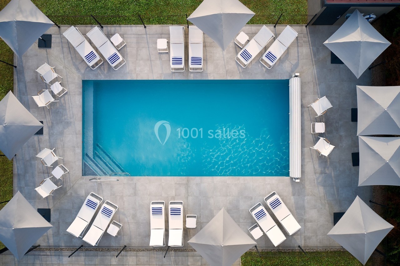 Vue aérienne d'une piscine rectangulaire entourée de chaises longues, de parasols blancs et d'une terrasse en dalles grises.