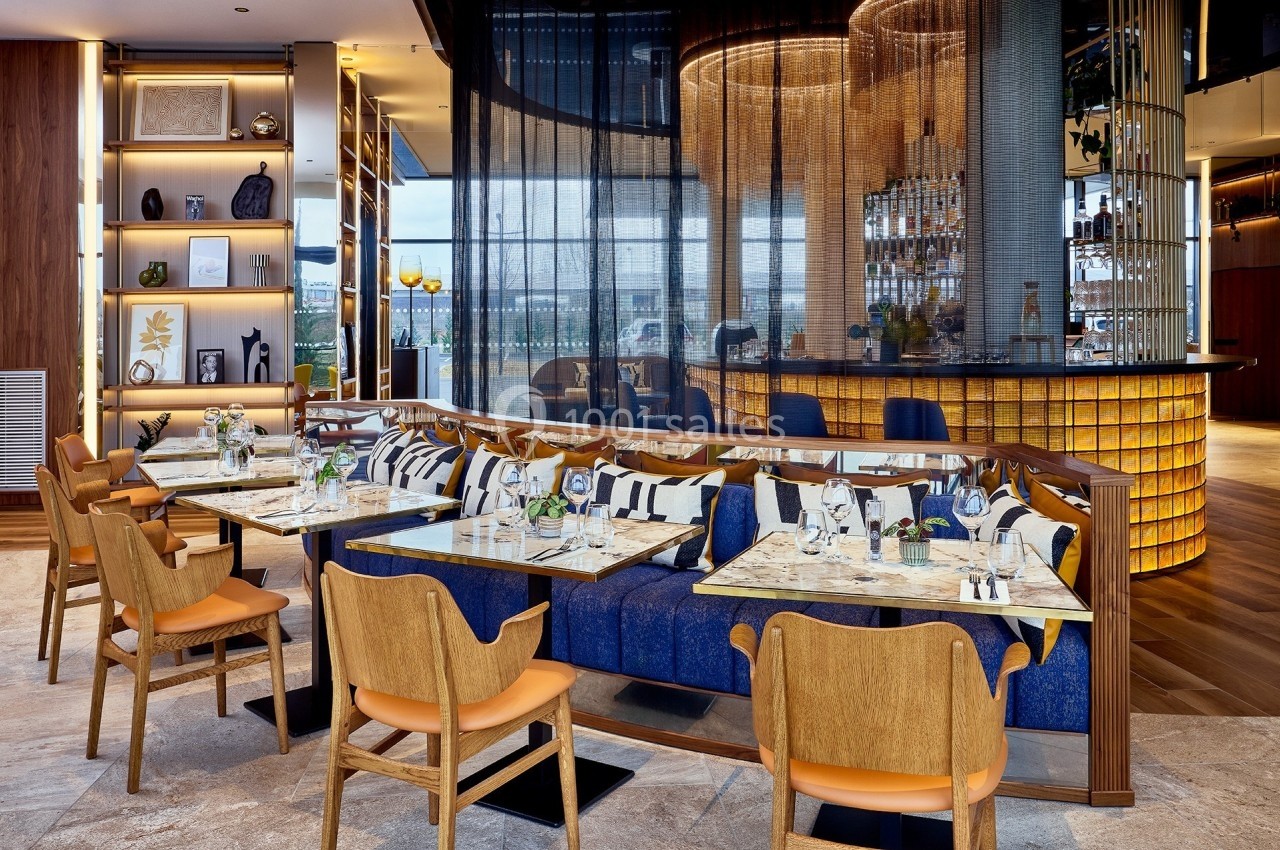 Salle de restaurant moderne avec tables dressées, chaises en bois, banquettes bleues et bar éclairé en arrière-plan.