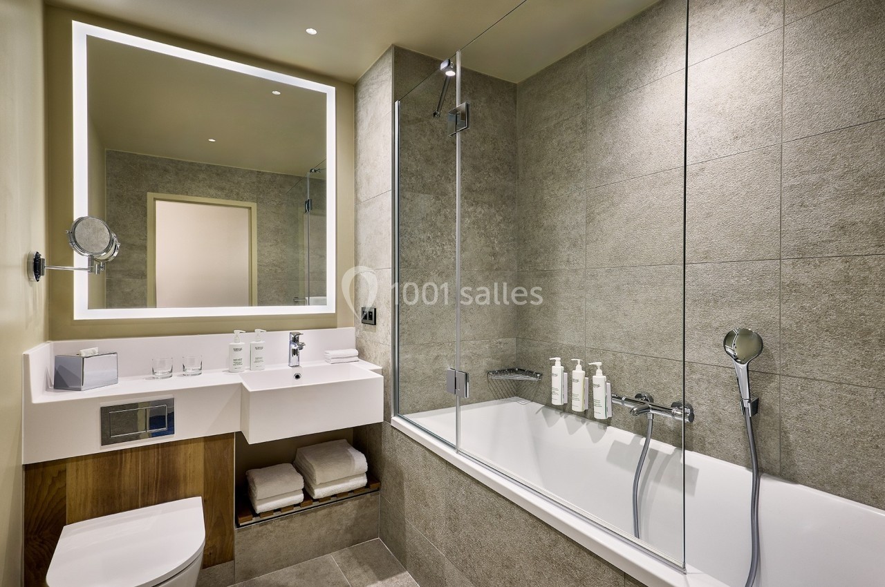 Salle de bain moderne avec baignoire, paroi vitrée, lavabo, miroir éclairé et étagère contenant des serviettes pliées.