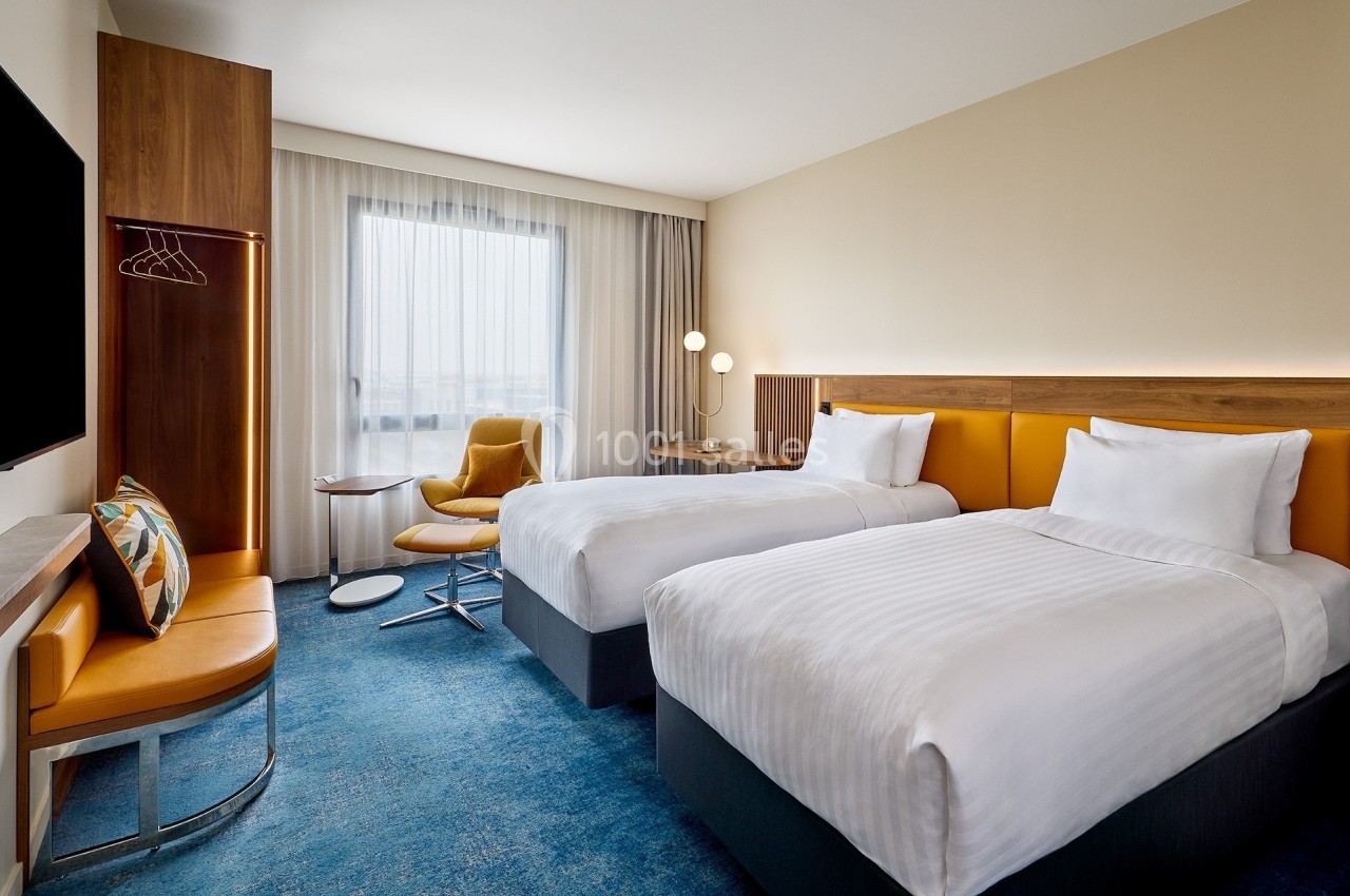 Chambre d'hôtel moderne avec deux lits simples, fauteuil jaune, bureau, et grande fenêtre laissant entrer la lumière…