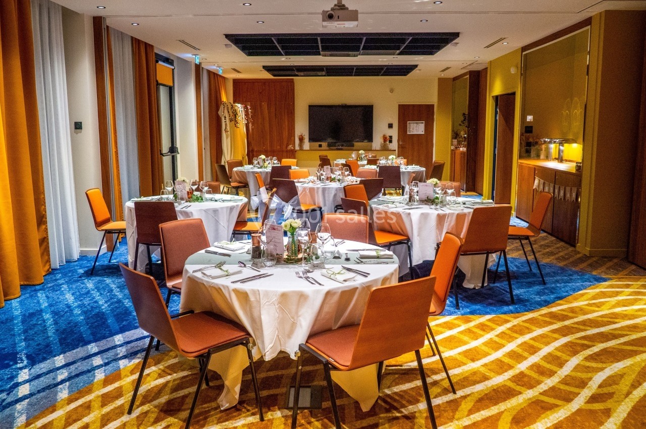 Salle de réception avec tables rondes dressées, chaises orange, éclairage chaleureux et décoration sobre.