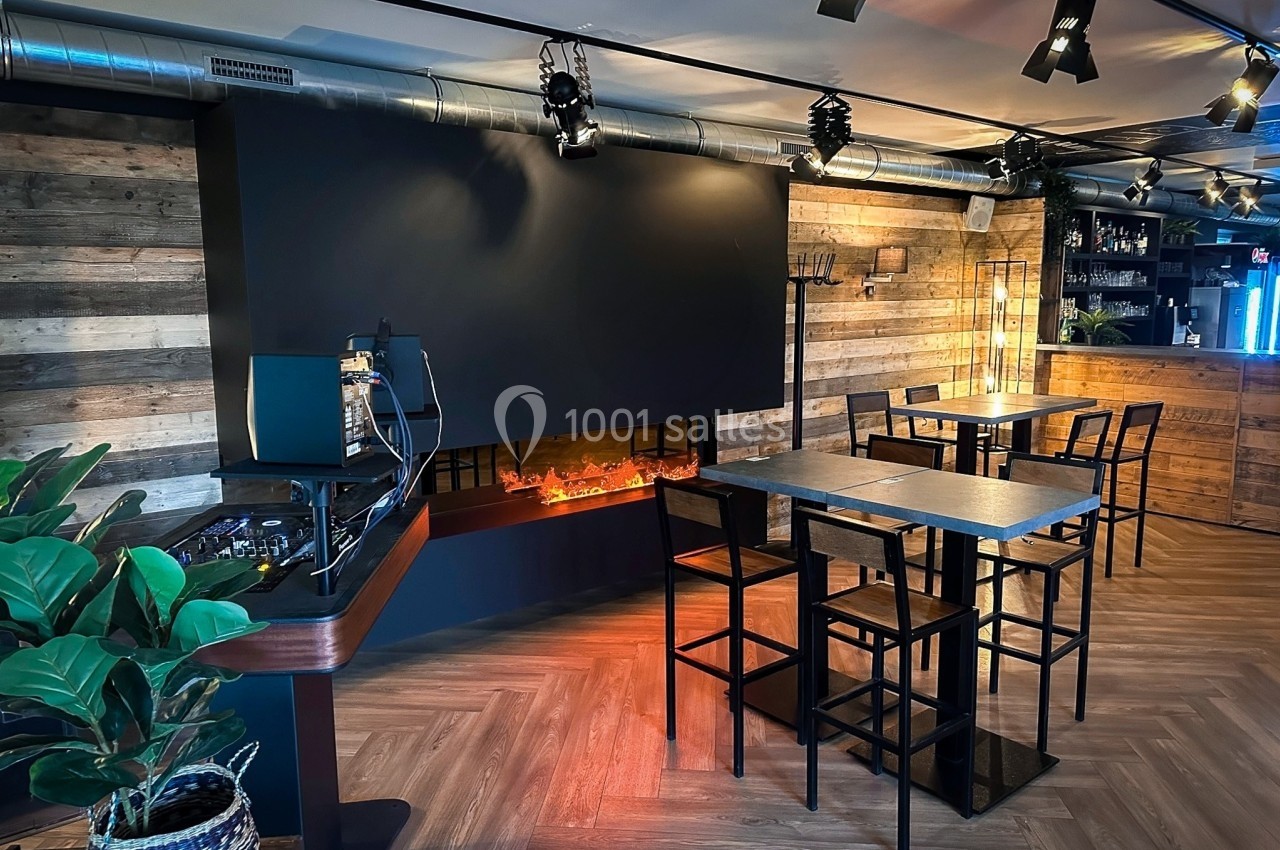 Salle moderne avec tables hautes, chaises noires, cheminée électrique et éclairage tamisé, décor en bois et métal.