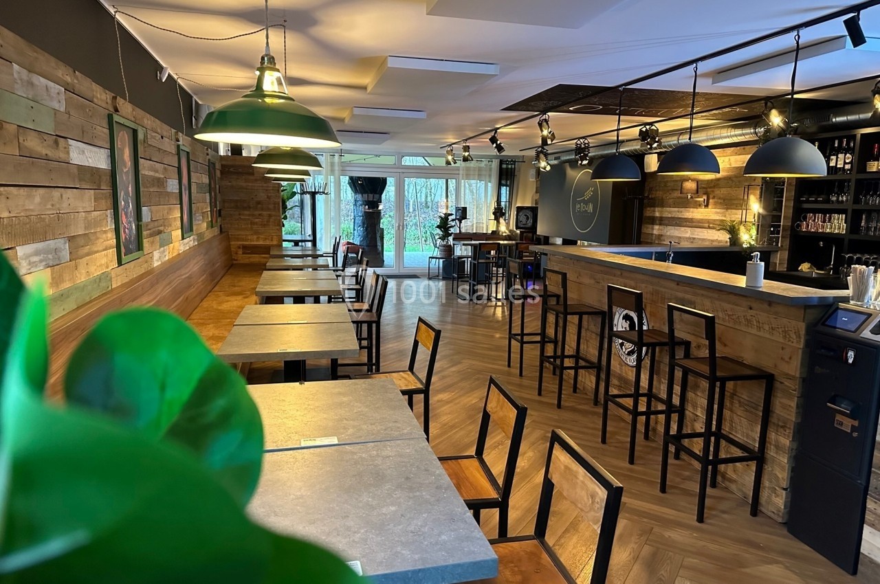 Salle de restaurant moderne avec tables en bois, chaises noires, éclairage suspendu et bar en arrière-plan.