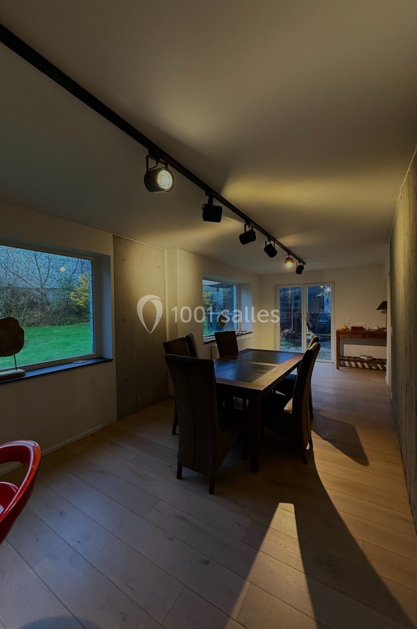 Salle à manger moderne avec table en bois, chaises sombres, éclairage sur rail et vue sur un jardin à travers de grandes…