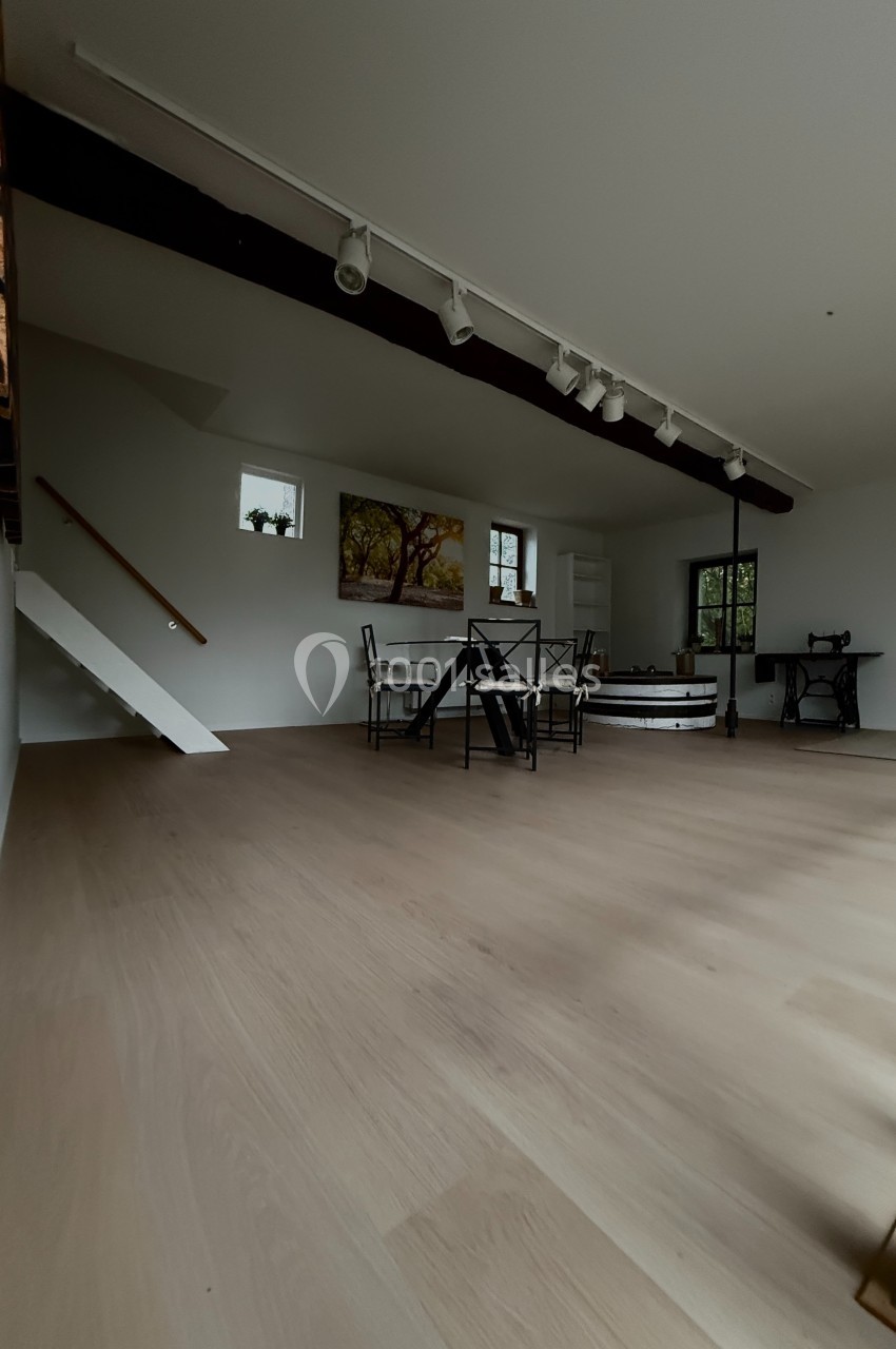 Salle lumineuse avec parquet clair, table et chaises au centre, escalier blanc et machine à coudre ancienne en arrière-plan.