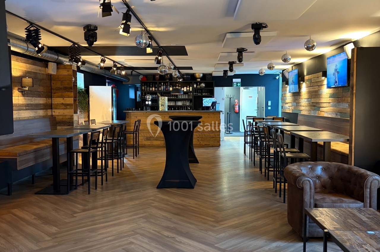 Salle moderne avec parquet, tables hautes, fauteuil en cuir, bar en bois et éclairage tamisé.