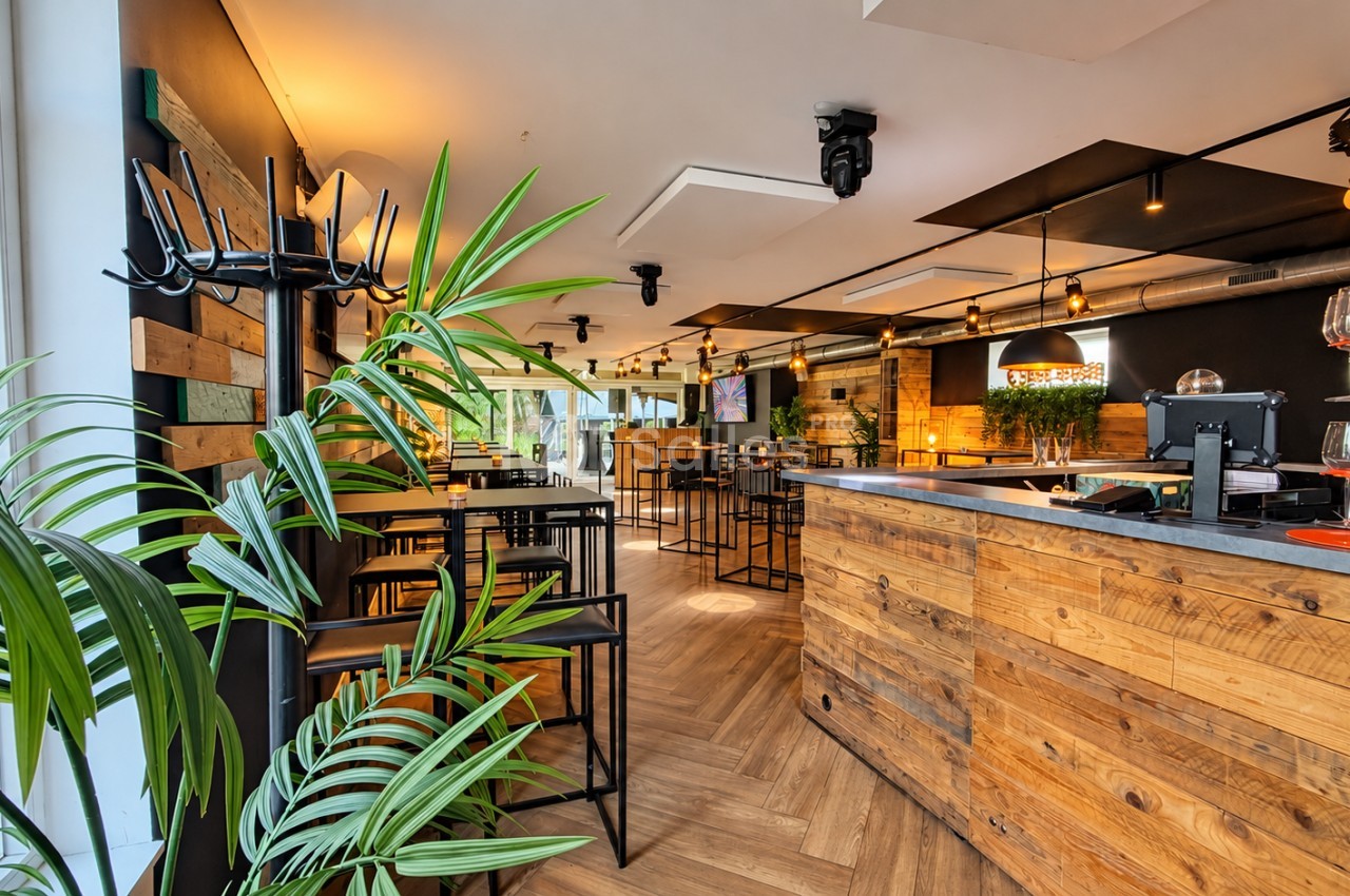 Intérieur de bar-restaurant au parquet en chevrons, avec un long comptoir en bois brut, des tables hautes noires, des plantes vertes et des suspensions industrielles.