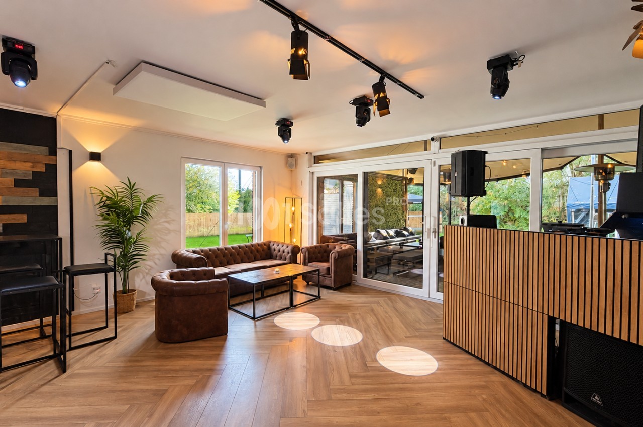 Salle à l'éclairage tamisé avec canapés en cuir brun capitonnés autour d'une table basse, console de DJ en bois sur la droite et grandes baies vitrées donnant sur un jardin.