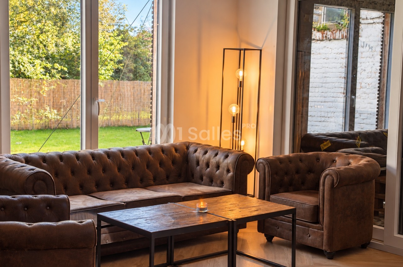 Salon avec canapé et fauteuils en cuir brun capitonné, table basse en bois et métal, lampadaire à ampoules apparentes, et grandes baies vitrées donnant sur un jardin.