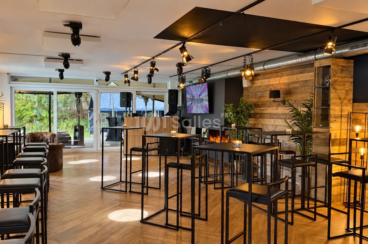 Salle de réception aménagée avec des tables hautes noires et tabourets, murs en bois de palettes, baies vitrées donnant sur un jardin et spots au plafond.