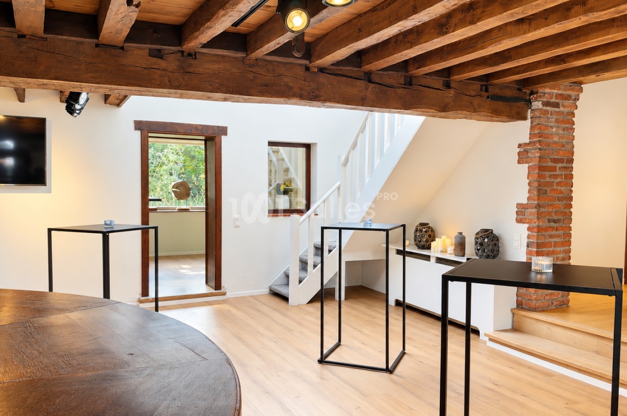 Pièce ouverte aux poutres de bois apparentes, avec un escalier blanc, une colonne en briques, une grande table en bois massif au premier plan et des tables hautes noires vides.