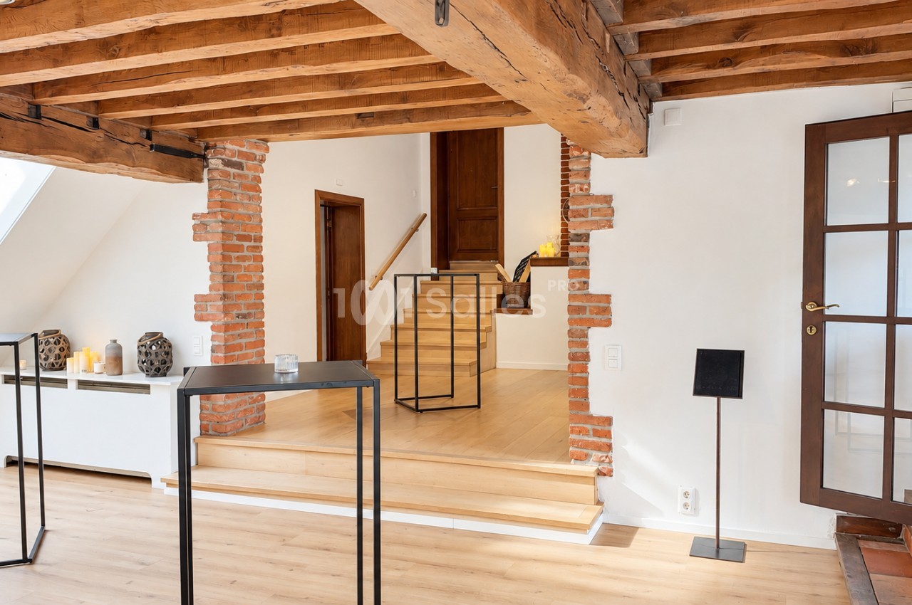 Pièce aux poutres de bois apparentes et murs blancs, avec des murs en briques, un escalier en bois à deux niveaux et un sol clair en parquet.