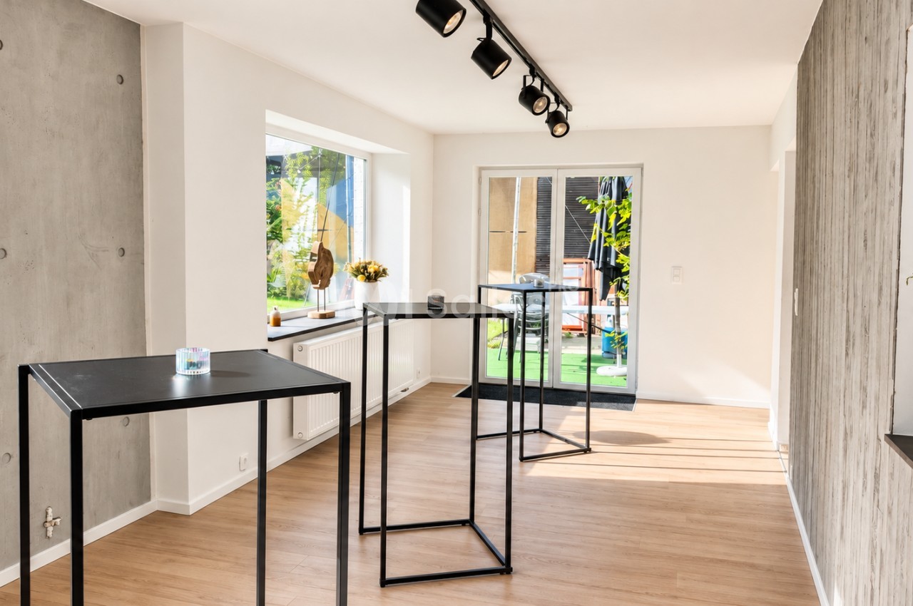 Pièce lumineuse aux murs blancs et sol en parquet clair, avec plusieurs tables hautes noires et une porte-fenêtre donnant sur un jardin.