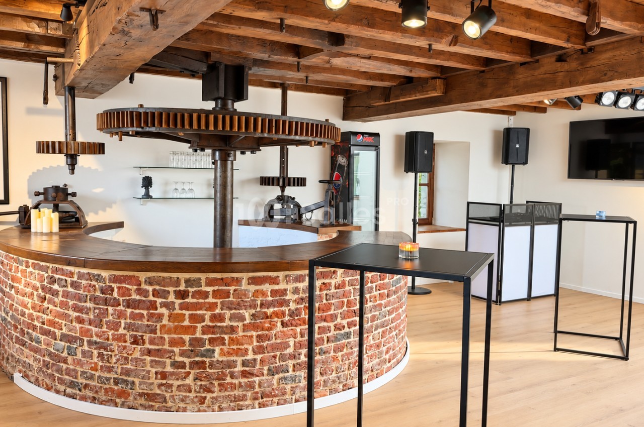 Bar circulaire en briques apparentes surmonté d'un plateau en bois, sous une charpente en poutres apparentes et de grands engrenages en bois, dans une salle aux murs blancs.