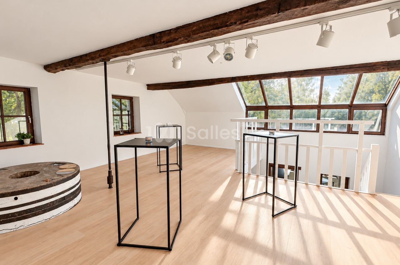Grande pièce sous combles au parquet clair, poutres apparentes, rails d'éclairage, hautes tables noires épurées et grande verrière donnant sur des arbres.