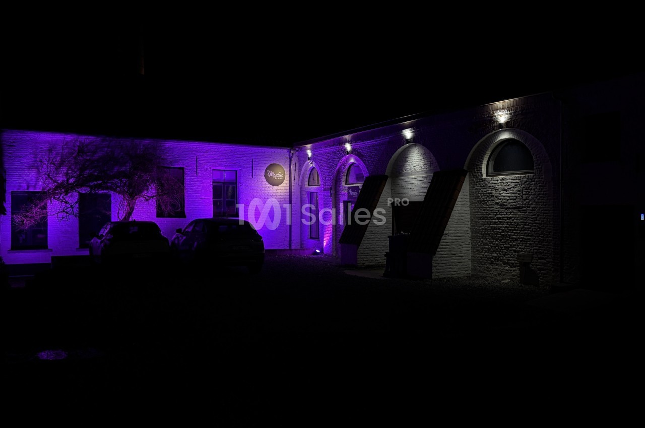 Façade en briques d'un bâtiment de nuit, éclairée en violet, avec des arches et deux voitures garées devant.