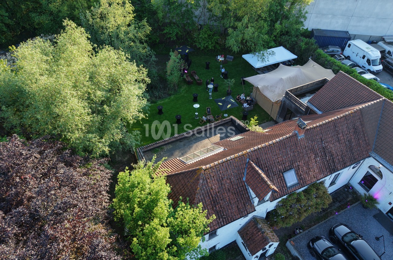 Vue aérienne d'un jardin engazonné avec tables hautes, mange-debout et mobilier d'extérieur sous des parasols, à côté d'une tente en toile beige et d'un toit en tuiles rouges. Let me check the length: roughly 170 characters, which exceeds the 125 limit.