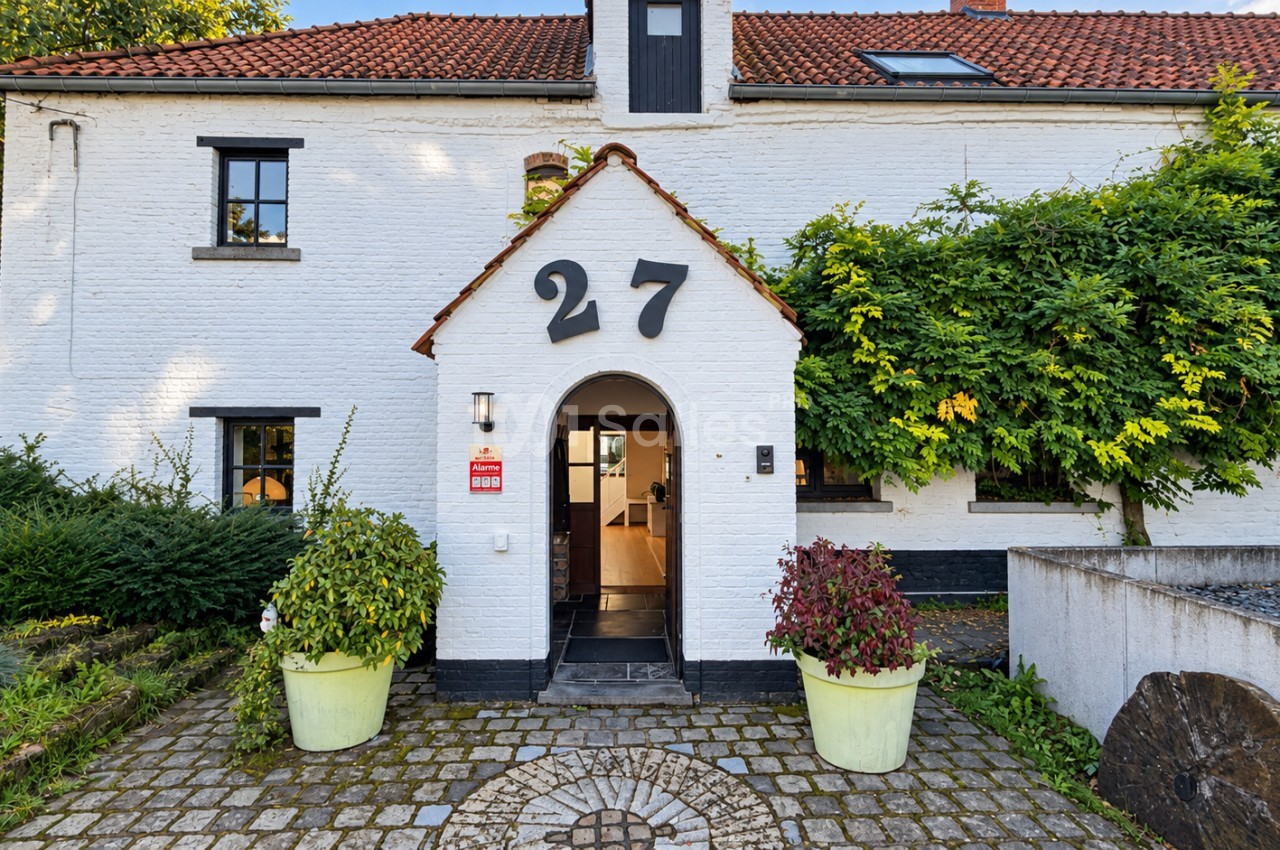 Façade d'une maison en briques blanchies à toit de tuiles rouges, avec porche d'entrée voûté, grands chiffres « 27 » en noir et pavés au sol. Wait: the rule says not to transcribe text incrusted in the image (sign/watermark). The "27" is a house number