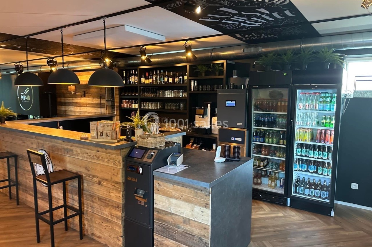 Comptoir en bois dans un espace moderne avec étagères de bouteilles, réfrigérateurs de boissons et éclairage suspendu.