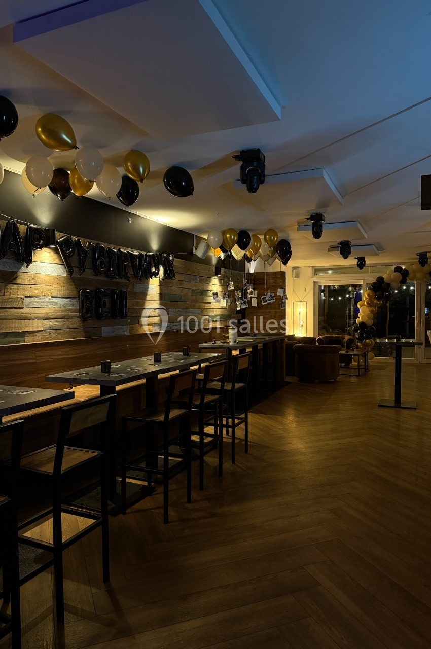Salle décorée avec des ballons noirs, dorés et blancs, tables hautes en bois et éclairage tamisé.