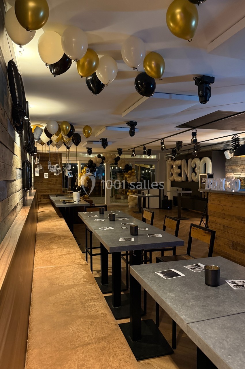 Salle décorée avec des ballons dorés, noirs et blancs, tables alignées et ambiance festive dans un espace moderne.