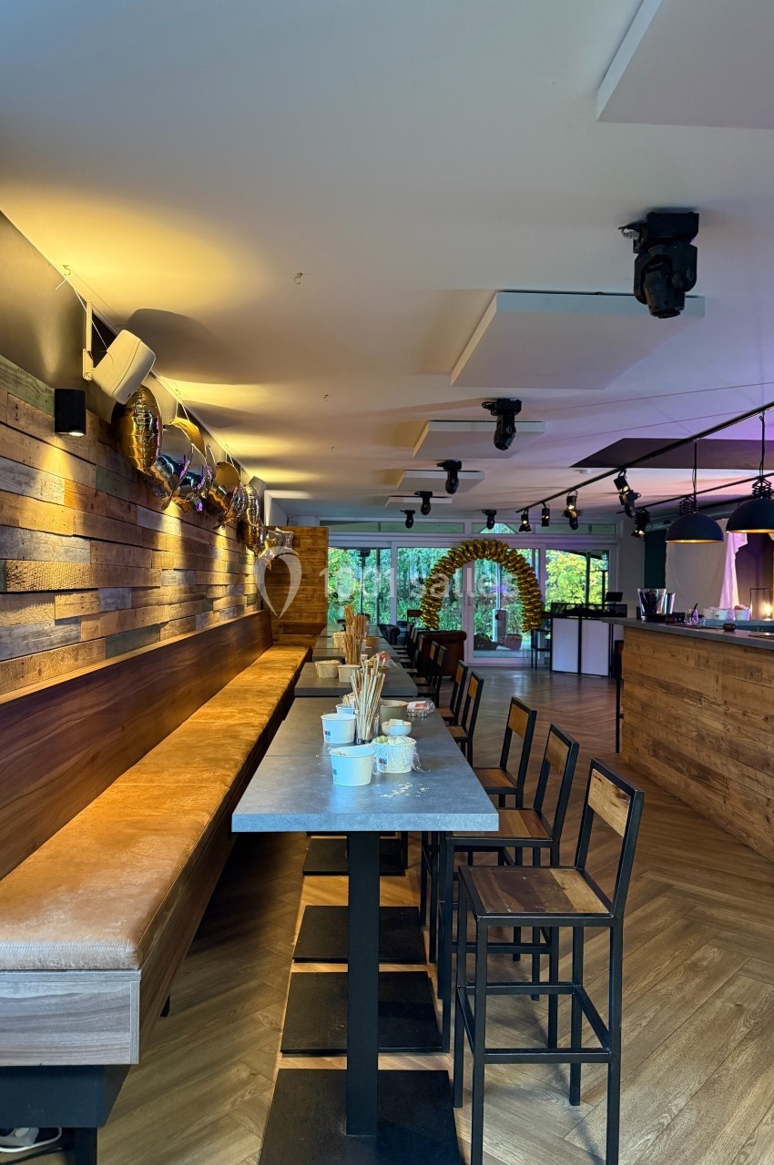 Salle moderne avec tables hautes, chaises noires, éclairage chaleureux et vue sur un extérieur verdoyant.