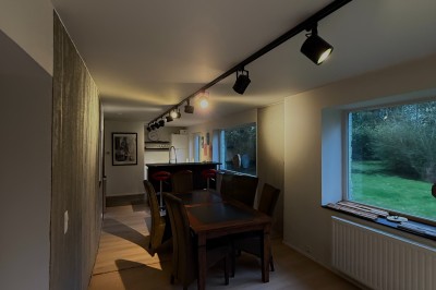 Salle lumineuse avec parquet clair, table et chaises au centre, escalier blanc, machine à coudre ancienne et tableau au mur.