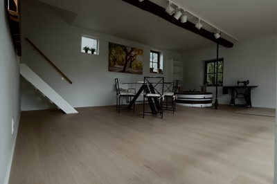 Salle lumineuse avec parquet clair, table et chaises au centre, escalier blanc, machine à coudre ancienne et tableau au mur.