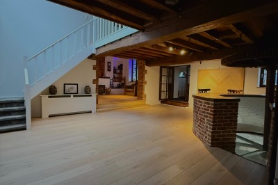 Salle lumineuse avec parquet clair, table et chaises au centre, escalier blanc, machine à coudre ancienne et tableau au mur.
