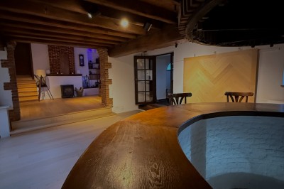 Salle lumineuse avec parquet clair, table et chaises au centre, escalier blanc, machine à coudre ancienne et tableau au mur.