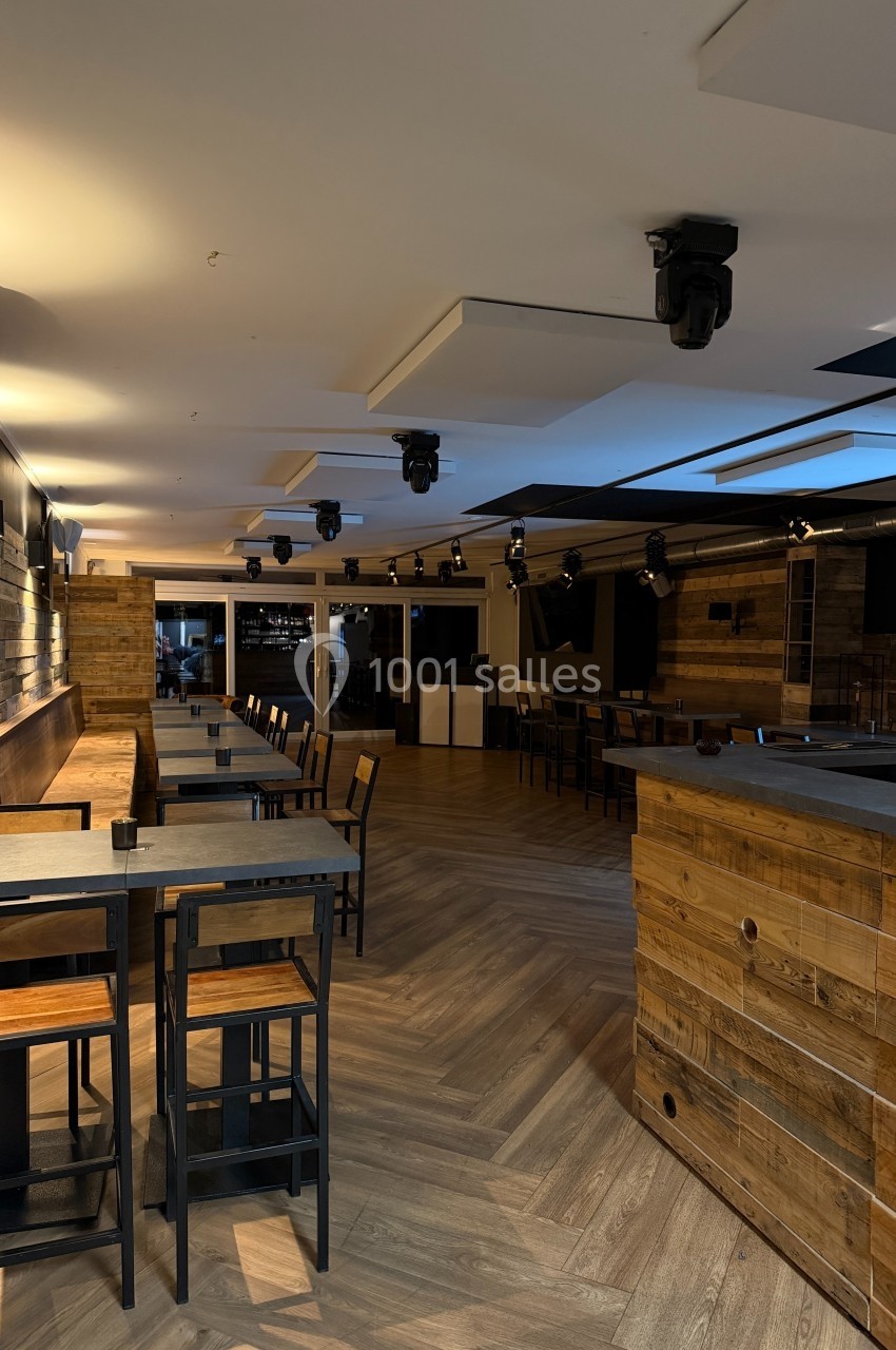 Salle moderne avec parquet, tables hautes, murs en bois et éclairage tamisé, aménagée pour des événements ou repas.