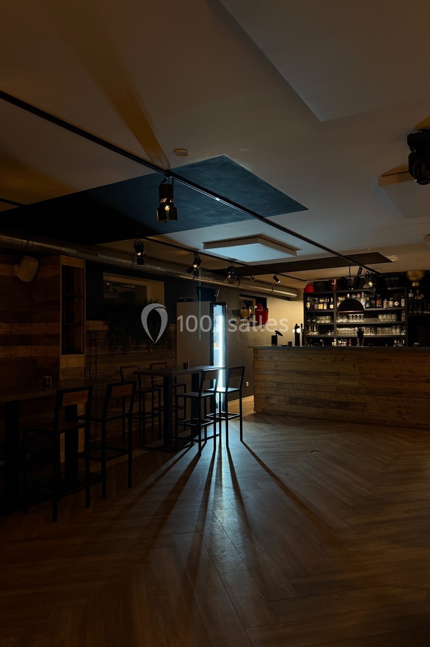 Salle de bar sombre avec comptoir en bois, tables hautes et lumière naturelle entrant par une porte vitrée.