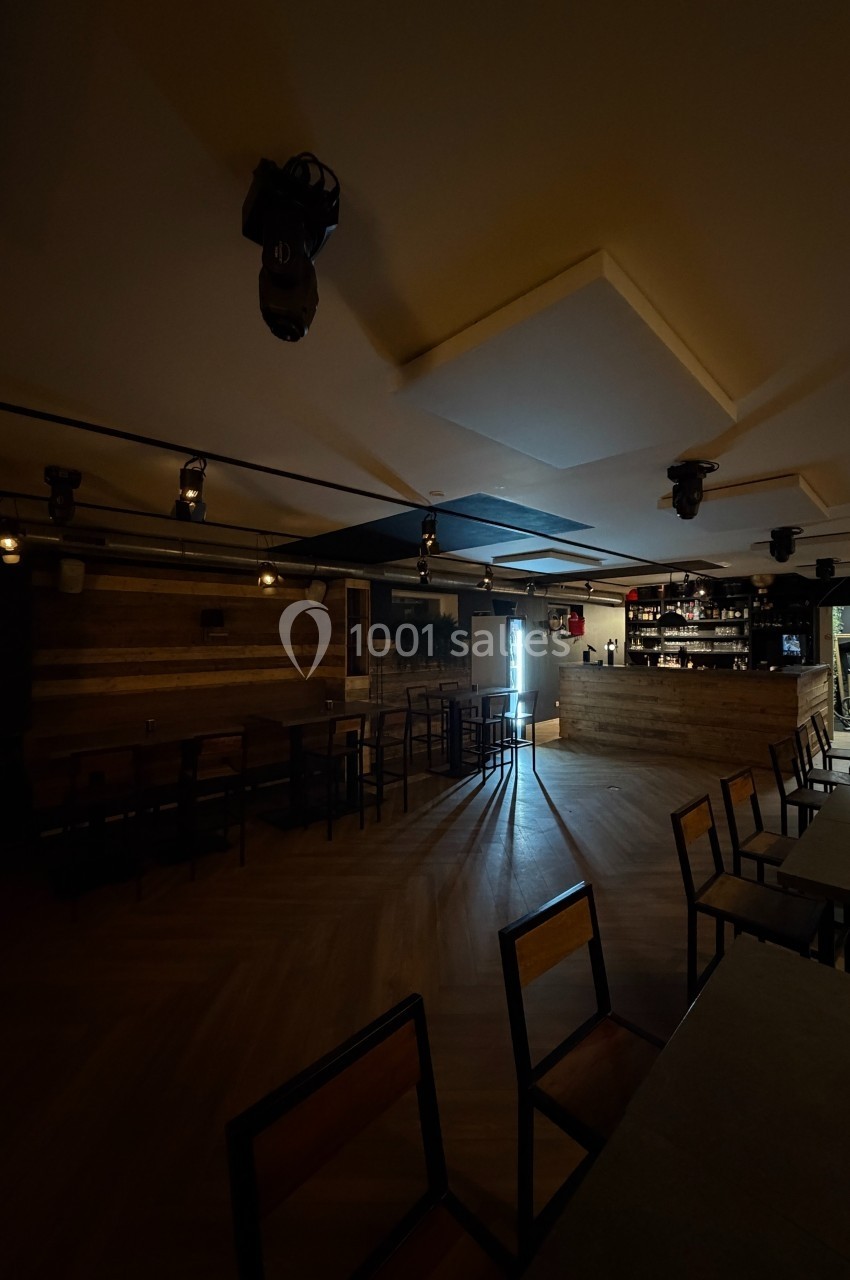 Salle sombre avec tables et chaises en bois, éclairée par une lumière naturelle provenant d'une porte ouverte.