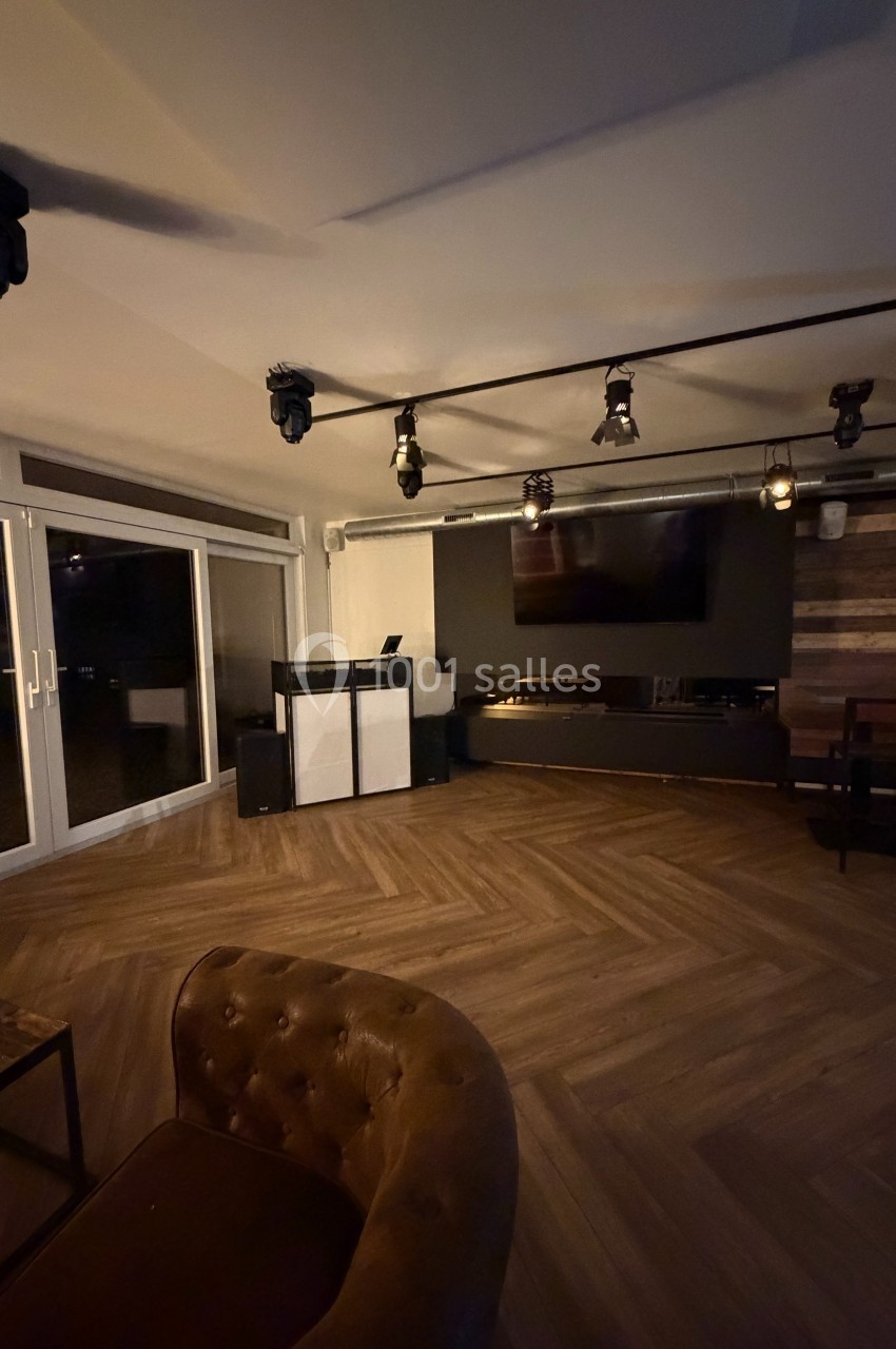 Salle moderne avec parquet en chevrons, murs en bois, éclairage suspendu, écran mural et grandes baies vitrées.